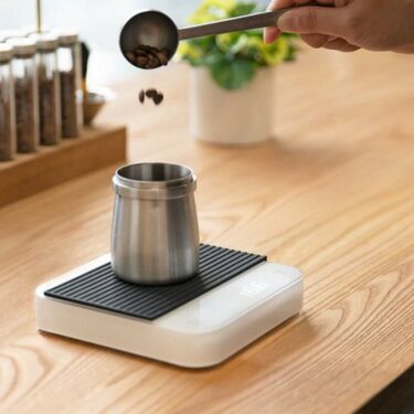 Grãos de café são depositados num recipiente/copo de metal que se encontra em cima de uma balança branca. No fundo há uma planta verde num vaso branco, em cima da mesma mesa de madeira.