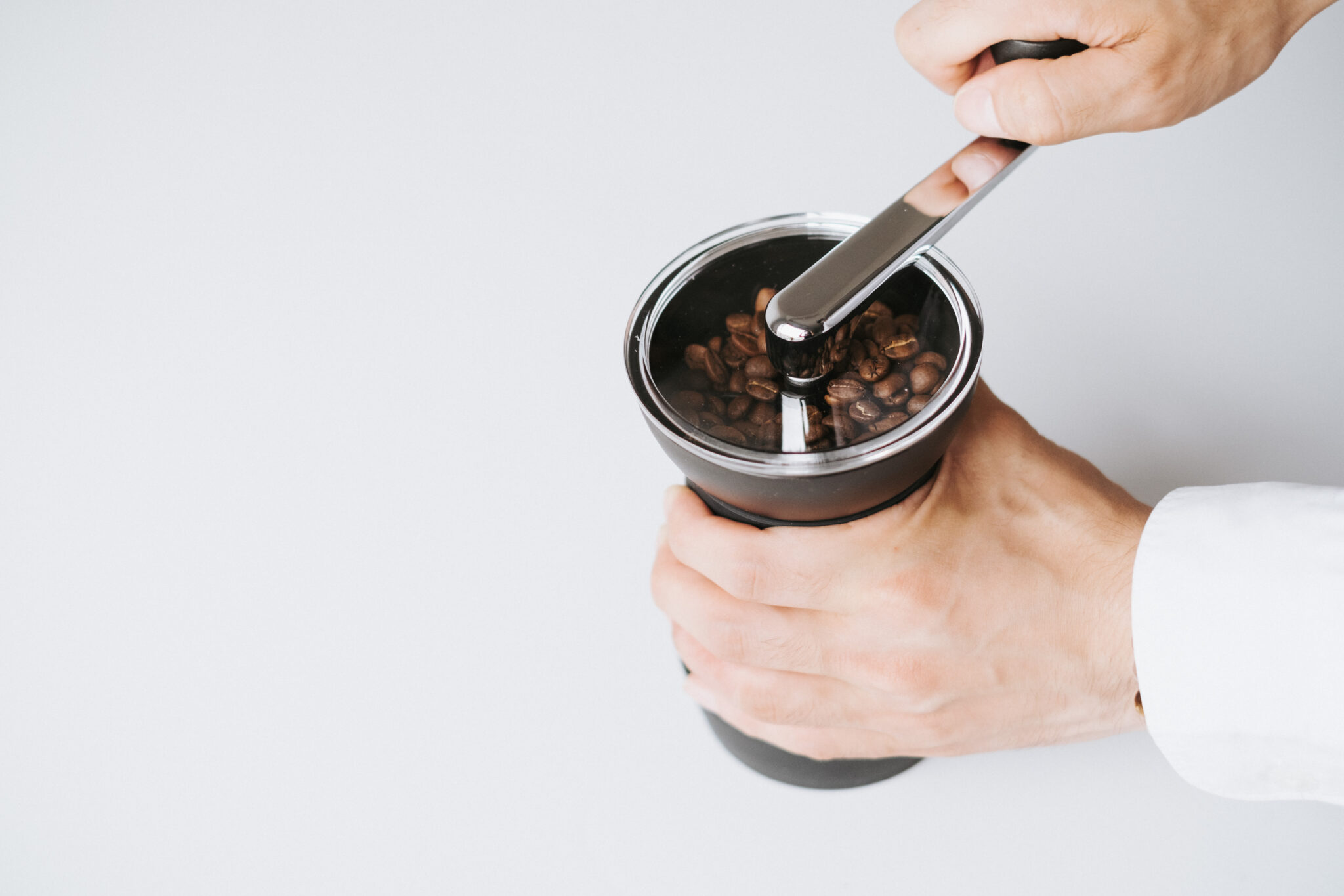 Moinho Skerton Pro 3 Duas mãos a segurar o moinho Skerto Pro da Hario enquanto moem os grãos castanhos de café que se encontram dentro do moinho.