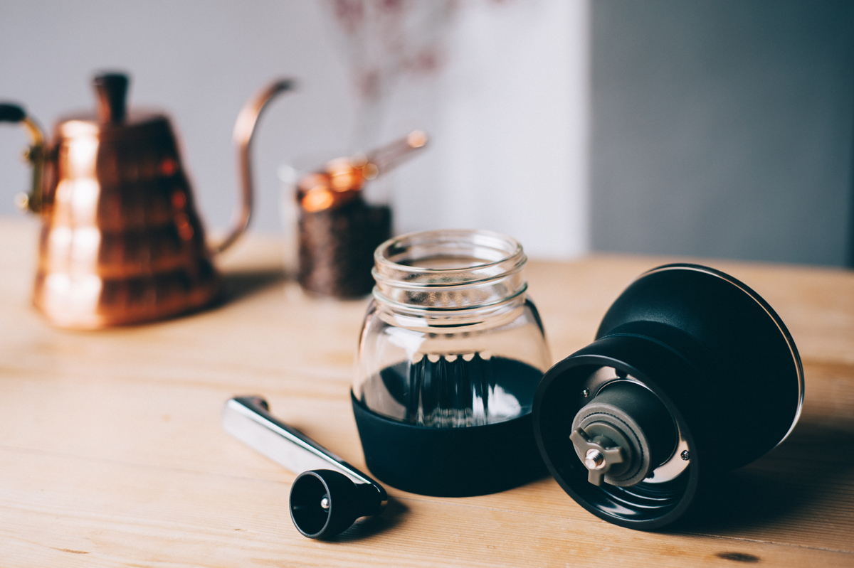 Moinho Skerton Pro 4 Moinho Skerton Pro da Hario desmontado com chaleira e solher de cobre no fundo. Em cima de uma mesa de madeira.